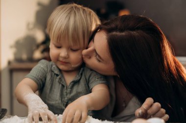 Bir annenin bebeğinin yanağını öptüğü samimi bir sahne mutfak masasında unla oynarken yavaş ebeveynlik anlarının şefkatli duygusal sıcaklığını ve günlük güzelliğini vurguluyor.