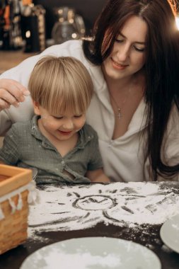 Neşeli bir anne ve bebek, unla oynarken ve mutfak masasına güneş çizerken birbirlerine bağlanıyorlar. Bağlantının, yaratıcılığın ve günlük aile hayatının iç açıcı bir sahnesini yaratıyorlar.