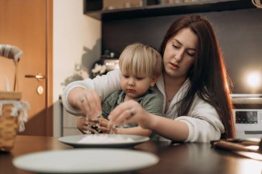 Küçük çocuk un karıştırırken anne ona sevgi dolu gerçek bir hayat sunuyor dokunsal keşiflerle dolu bir an evde sıcak bir mutfak ortamında neşe ve yakın ebeveyn bağı kuruyor.