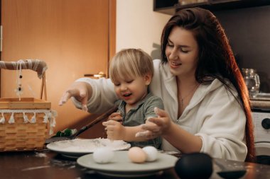 Gülümseyen çocuk un ve yumurtalarla oynarken anne de ellerini ılık sevgi ve bağ ile dolu sıcak mutfak ortamında neşeli bir duyusal öğrenme deneyimi yaratıyor.