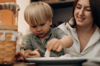 Meraklı bir çocuk tabakta un karıştırırken annesi sevgi dolu bir şekilde yanında yaratıcı keşif ve duyusal öğrenme anını izler. Samimi bir ev mutfağında.