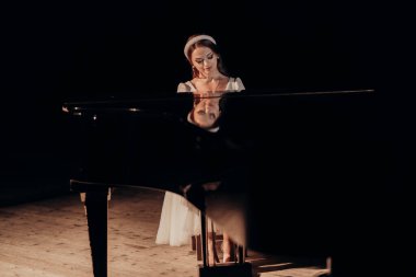 Beyaz elbiseli bir piyanist zarif bir şekilde kuyruklu bir piyanonun başında oturuyor, onun sakin ifadesi enstrümanın parlak yüzeyine yansıyor. Spot ışıkları sonsuz bir ortam yaratır.