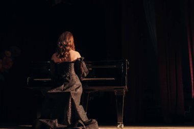 Göz kamaştırıcı siyah elbisesi olan bir kadın kuyruklu piyanonun başında oturur ve sırtını kameraya çevirir. Loş sahne ışıkları onun siluetini ve sahnenin zarafetini vurguluyor..