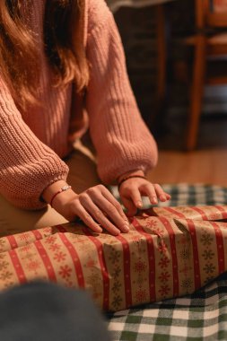 Tatillerde hediye paketi yapan rahat bir süveter giyen biri hediyeyi kareli bir battaniyeye oturmuş bayram kâğıdıyla dikkatlice paketliyor. İçerideki sıcak ortam tatil atmosferini çağrıştırıyor..