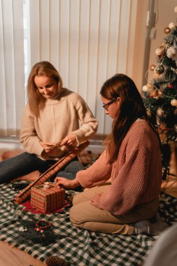 Kareli battaniyeye oturmuş, Noel için hediyeler paketleyen iki kadın. Bir kadın hediyesine kurdele bağlarken diğeri de yakındaki süslemeleri yerleştiriyor. Süslü bir Noel ağacı