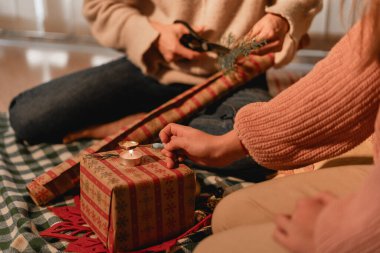 İki insan hediye paketleri, ellerinde makas ve sicim ve yeşillik gibi dekoratif unsurlarla bir hediye paketliyor. Kareli battaniyeyle iç mekan ayarları.