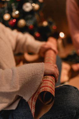 Konforlu Noel Hediyesi Paketleme Sahnesi elinde bir rulo ambalaj kağıdıyla dekore edilmiş bir Noel ağacının yanında, yumuşak ışıklandırmalı bir şekilde oturan birinin yakın çekimi. Sahne sıcak bir tatil atmosferini çağrıştırıyor.