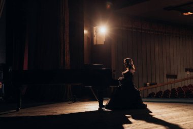 Siyah elbiseli bir kadın boş bir sahnede kuyruklu piyano çalar. Dramatik ışıklandırma ve gölgeler konser salonunda büyüleyici ve sinematik bir atmosfer yaratıyor.