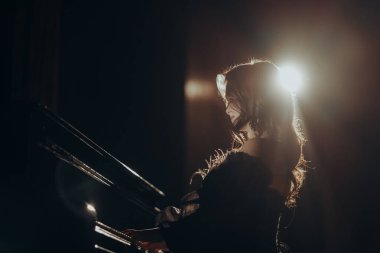 Parlak bir spot ışığı altında piyano çalan bir kadının dramatik silueti. Onun zarif profili ve dalgalı saçı bu karamsar, sanatsal kompozisyona sinematik bir dokunuş katıyor.