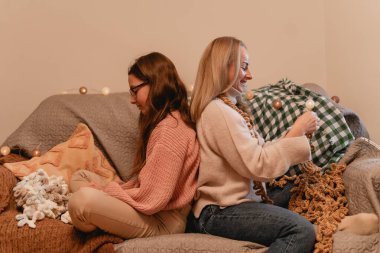 Bir kadın ve bir kızın arka arkaya kanepede oturup el sanatları yaptığı samimi bir sahne. Oda yumuşak ışıklandırma ve battaniyelerle dekore edilmiş, sıcak bir atmosfer yaratıyor..