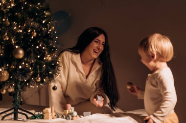 Anne ve çocuk neşeli bir bağı paylaşıyor. Parlayan ışıklarla neşeli bir ağacın yanında oynuyorlar. Özel kış kutlamalarında sıcak ve şefkatli bir ev akşamını yakalıyorlar.