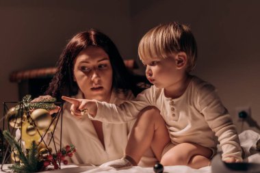 Bir anne ve küçük çocuğu sıcak ve yumuşak bir odada birlikte oturuyorlar. Anne, süslemelerin dekoratif düzenini gösteren çocuğa dikkatle bakar..