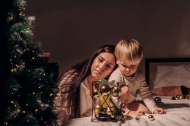 Anne ve çocuk başlarını yan yana oturarak sakin ve sakin bir ışıkta şenlikli dekorasyonlara hayran kalarak bir anlık yakınlık yansıması ve barışçıl bir bağ paylaşıyorlar.