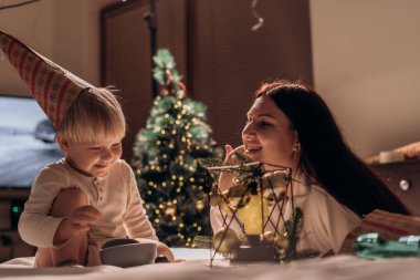 Neşeli bir anne ve küçük oğlu evde kutlama yapıyorlar. Çocuk bir parti şapkası takar ve bir kâseyle oynar, bu arada anne onunla gülümser ve etkileşim kurar. Süslü bir Noel ağacı...