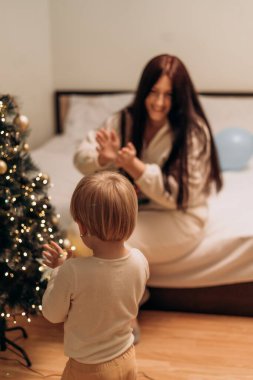 Noel Ağacı 'na yakın neşeli bir aile kutlaması bir kadın ve bir çocuğun Noel ağacının yanında yumuşak ışıklandırması ve rahat bir atmosferle kutlama sahnesi. Kadın, çocukla şakalaşıyor.
