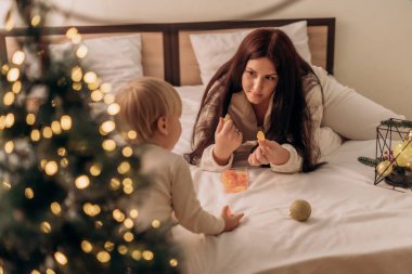 Bir anne ve küçük çocuğu bir yatakta oturuyorlar, neşeli bir andalar. Çocuk ona merakla bakarken, anne renkli şekerlemeler tutuyor. Sahne sıcacık, dekore edilmiş