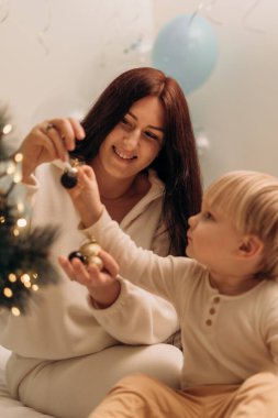 Neşeli bir anne ve küçük oğlu birlikte bir Noel ağacı süslüyorlar. Çocuk başka bir süs eşyası ararken anne elinde bir süs taşırken gülümsüyor. Yumuşak ışıklandırma ve şenlikli dekorasyon