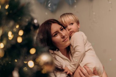 Bir anne çocuğunu yanında tutarken etrafını Noel süsleriyle sarmış düşünceli bir şekilde bakar. Yumuşak ışık sıcak ve rahat bir atmosfer yaratır. Anne Çocuk Konforlu Noel Ayarlarını Kucaklıyor