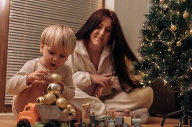 Genç bir çocuk altın Noel süslerini istiflerken bir kadın onu gülümseyerek izliyor. Sahne, süslü bir Noel ağacı ve yumuşak ışıklandırmayla sıcak bir tatil ortamı yaratıyor..