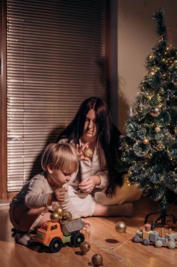 Noel ağacını süsleyen bir anne ve çocuk. Anne arka planda yardımcı olurken, çocuk bir oyuncak kamyon ve altın süslemelerle oynuyor. Sıcak ışıklandırma sıcak bir tatil ortamı yaratır..