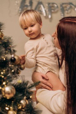 Bir anne çocuğunu Noel ağacının yanında nazikçe tutar. Onların yakınlığı, sevgi ve ilgiyi, tatillerde içtenlik, güven ve duygusal bağlarla dolu sakin bir anda yansıtır.