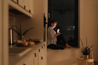 Geceleri telefon kullanan bir kadın karanlık mutfakta pencerenin önünde otururken evde sakin ama izole edici modern akşam rutininin bir parçası olarak ekran bağımlılığını dijital yalnızlığı ve mavi ışığa maruz kalma gösteriyor.