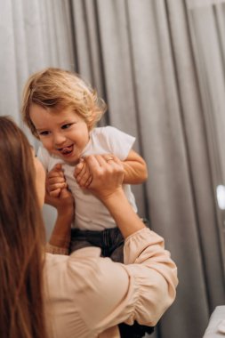 Genç anne, gülümseyen çocuğu kaldırarak sevgi dolu bir ebeveyn sahnesi yaratıyor ve göz temasını, güveni ve birbirine bağlı duyguları paylaşarak gündelik mutluluğu paylaşıyor.