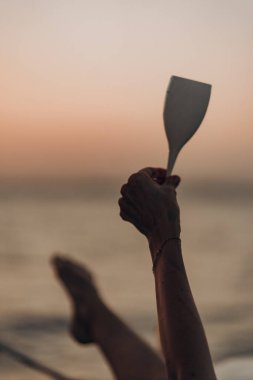 Günbatımına doğru bir spatula kaldıran bir kadının sanatsal yakın çekimi yatta uzanırken arka planda hafif bacaklar hayal ötesi kontrast karışımı bir tatil uysallığı ve yaratıcı detay yaratır.