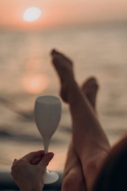 Altın saatinde beyaz kadehli kadınların eli zarif bir şekilde kapanıyor. Bacakları arka planda bulanık. Gün batımı renkleri sakin bir yolculuk ve rüya gibi bir sahil akşamında huzurlu bir lüks taşıyor.