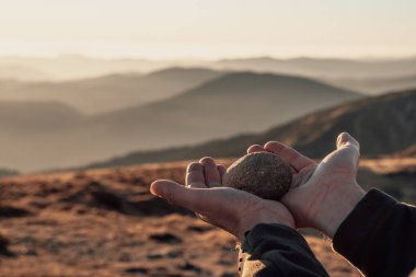 Sıcak akşam güneşiyle yıkanmış, yumuşak ve bulanık bir dağ zeminiyle, tek bir pürüzsüz taşı tutan eller, barışın farkındalığının basit ama güçlü bir görüntüsünü yaratıyor.
