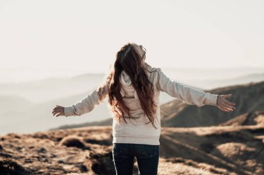 Genç bir kadın, kolları açık, güneşli bir dağ sırtında duruyor. Özgürlüğü ve doğayla bağlantıyı kucaklıyor. Temiz havanın ve dağlık arazinin nefes kesici panoramik manzarasının tadını çıkarıyor.