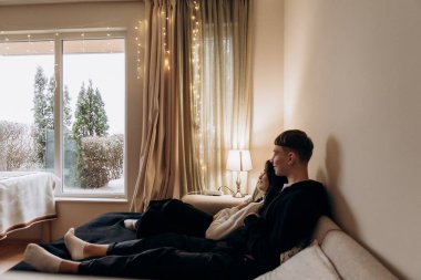 Genç çift kış gününü sessiz bir şekilde pencerenin içinde karlı bahçeleri seyrederek geçiriyorlar. Yumuşak ışık ve şenlikli bir tatil evi. Sıcak, huzurlu, sevgi dolu bir ortam.