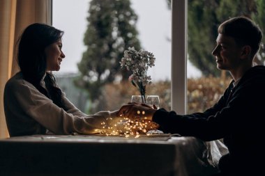 Romantik kış akşamının tadını çıkaran genç bir çift, şarap kadehleri ve şişe ılık Bokeh ışıkları eşliğinde sıcak bir masada el ele tutuşarak büyülü bir cazibe ve özel bir atmosfer ekliyor.