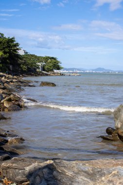 Güneşli bir günde sakin mavi sular, demirli tekneler ve doğal kaya oluşumlarıyla çekilen huzurlu bir plaj sahnesi. Güneş ışığı kıyı şeridinin güzelliğini arttırır. Mükemmel bir huzur anı sunar ve deniz kıyısından kaçar..