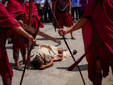 Dini giyinmiş bir adam büyük ahşap bir haç taşımak için mücadele eder ve düşer. Diğer muhafızlar önlerinde durup, kuyularını geçiyorlar.