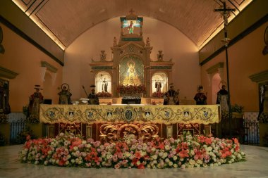 Dini heykeller ve canlı pembe ve turuncu çiçeklerle süslenmiş ayrıntılı, altın bir sunak bir kilise ortamında aydınlanıyor.