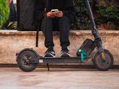 Elektrikli scooterlı bir bankta oturan ve elinde akıllı telefon tutan bir yemek dağıtıcısının yakın çekim görüntüsü. Siyah atletik giysiler ve spor ayakkabılar giyiyor. Modern ulaşım ile şehir sokak sahnesi