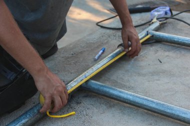 Elleri, beton bir yüzeye yerleştirilmiş metal boruları ölçmek ve işaretlemek için sarı bir mezura ve mavi bir işaret kullanılarak gösterilir.