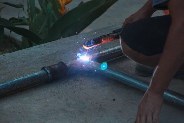 Metal tüpleri birleştirmek için çubuk kaynak makinesi kullanan bir insan açık havada gösteriliyor. Parlak kıvılcımlar ve parlayan kaynak havuzu üretiliyor.