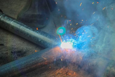 Yakın plan çekimde, Kalkan Metal Kaynak SMAW sırasında oluşan yoğun ışık, kıvılcımlar ve dumanlar, iki metal tüpü birleştirmek için kullanılan bir elektrot olarak gösterilir.