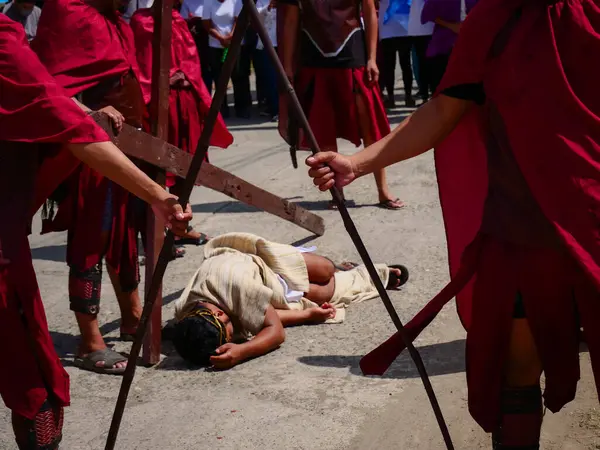 Dini giyinmiş bir adam büyük ahşap bir haç taşımak için mücadele eder ve düşer. Diğer muhafızlar önlerinde durup, kuyularını geçiyorlar.