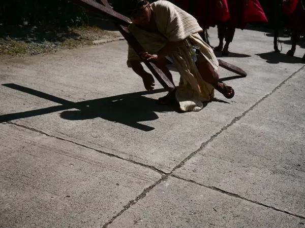 Dini giyinmiş bir adam kaldırımlı bir yol boyunca büyük bir ahşap haç taşımak için mücadele ediyor, uzun bir gölge bırakıyor. Diğer figürler takip ediyor