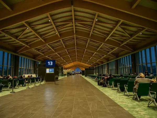 Yüksek ahşap kiriş tavanı ve geniş pencereleri olan geniş bir havaalanı bekleme alanı. Yeşil koltuklar ve 13 kapılı tabela görünüyor.