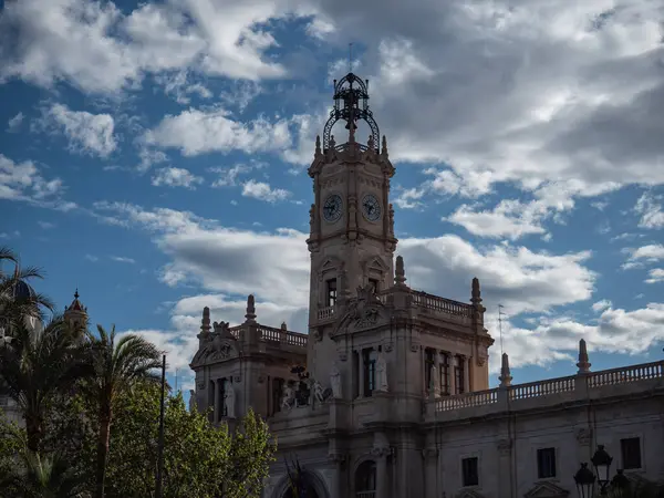 Detaylı bir görüntüde Valencia Belediye Binası 'nın süslü saat kulesi, dokulu bulutlar ve mavi parçalarla dolu dramatik bir gökyüzüne karşı yükseliyor. Sol alttaki palmiye ağaçları ve diğer yeşillikler görülebiliyor.
