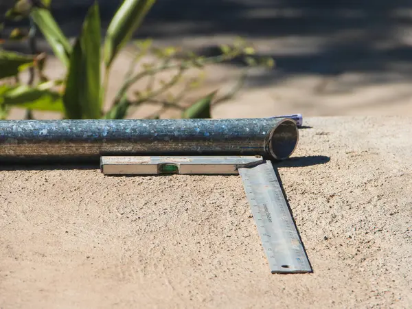 Silindirik metal bir tüp, küçük bir ruh seviyesi ve metal bir cetvelle birlikte sert bir beton yüzeyde bulunur. Tüpün arkasına mavi bir işaret yerleştiriliyor.