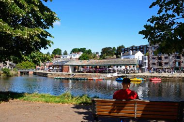 EXETER, UK - 22 AĞUSTOS 2023 - East Quay, Exeter, Devon, UK, Europe, 22 Ağustos 2023 'te Exe Nehri' nin karşısındaki dükkan ve restoranlara bakan bir bankta oturan adam.