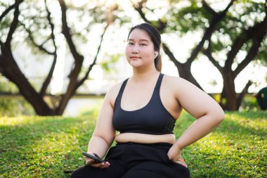 Dışarıda oturan bir kadının portresi, yeşillik ve doğal ışıkla çevrili, rahatlamış ve bir telefon tutuyor. Asyalı artı beden kadın.