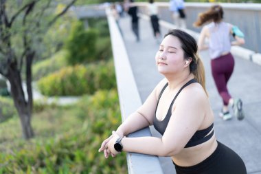 Bir kadının portresi, açık havada egzersiz yaptıktan sonra bir an rahatlarken, diğerleri yeşil bir manzaranın içinde koşuyor. artı boy kadın.