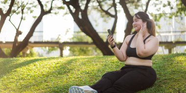 Bir kadının portresi çimlerin üzerinde oturur, güneşli bir parkta dinlenirken telefonuyla müziğin keyfini çıkarırken, rahatlık ve refahı somutlaştırır. Asyalı artı beden kadın.
