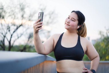 Spor kıyafetli gülümseyen bir kadının portresi dışarıda selfie çeker, şehir arka planında egzersiz ve doğanın tadını çıkarır. artı boy kadın.